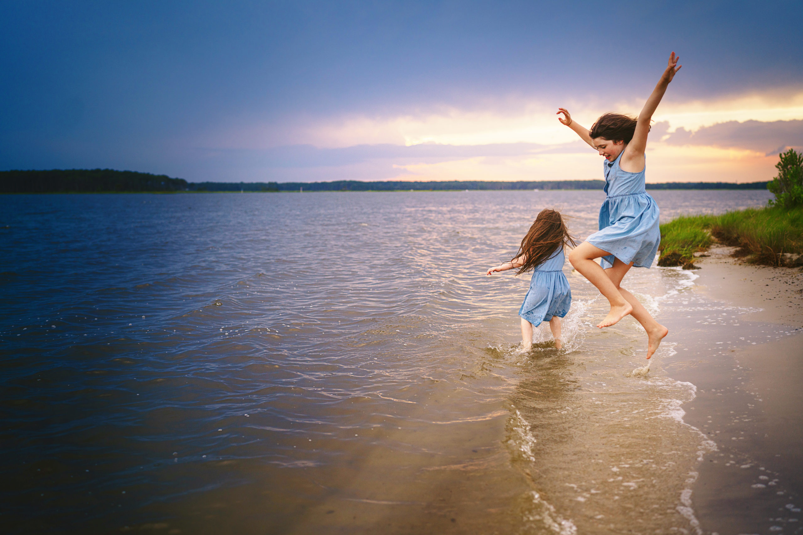 two young girls playing in the water at the beach of a lake during a sun set