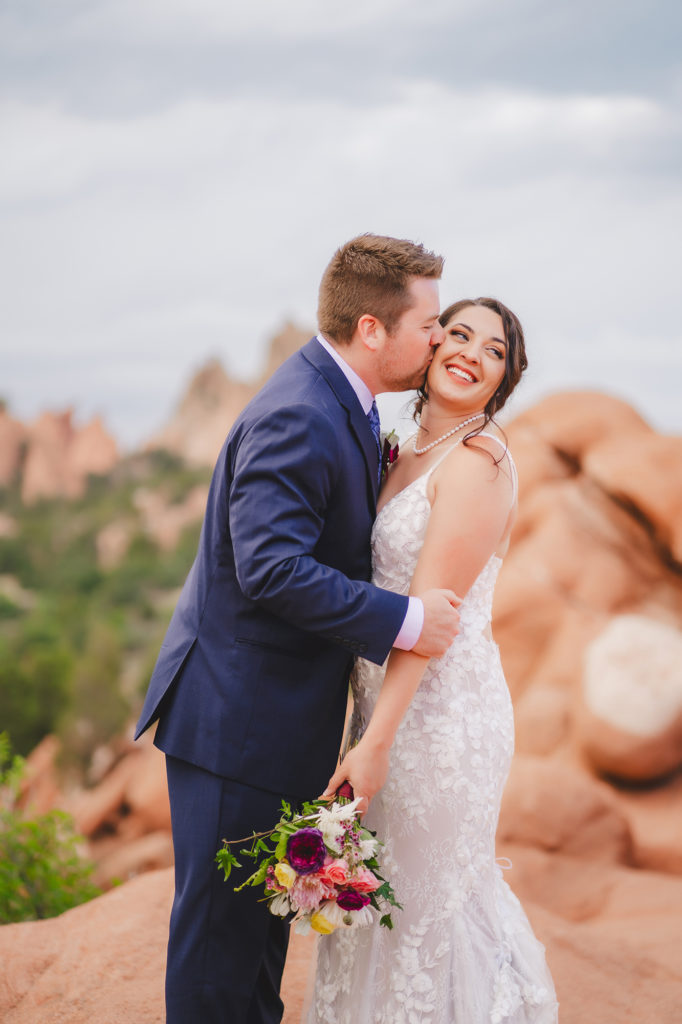 groom holding his brides arms and kissing her on the cheek as she smiles for their colorado elopement in garden of the gods