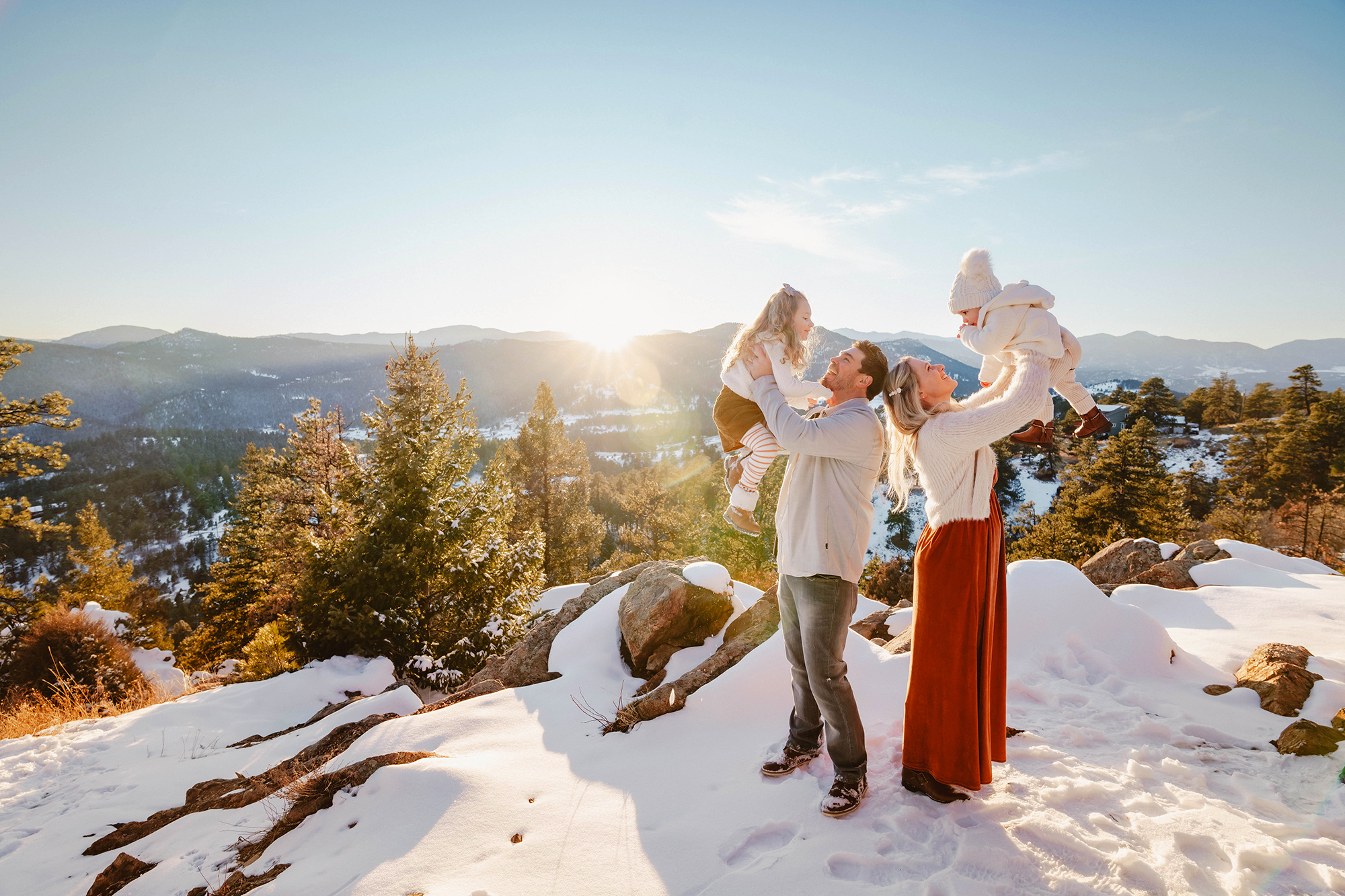 Parents lifting their children in the air on a snowy mountaintop, showing playful winter outfits for family photos.