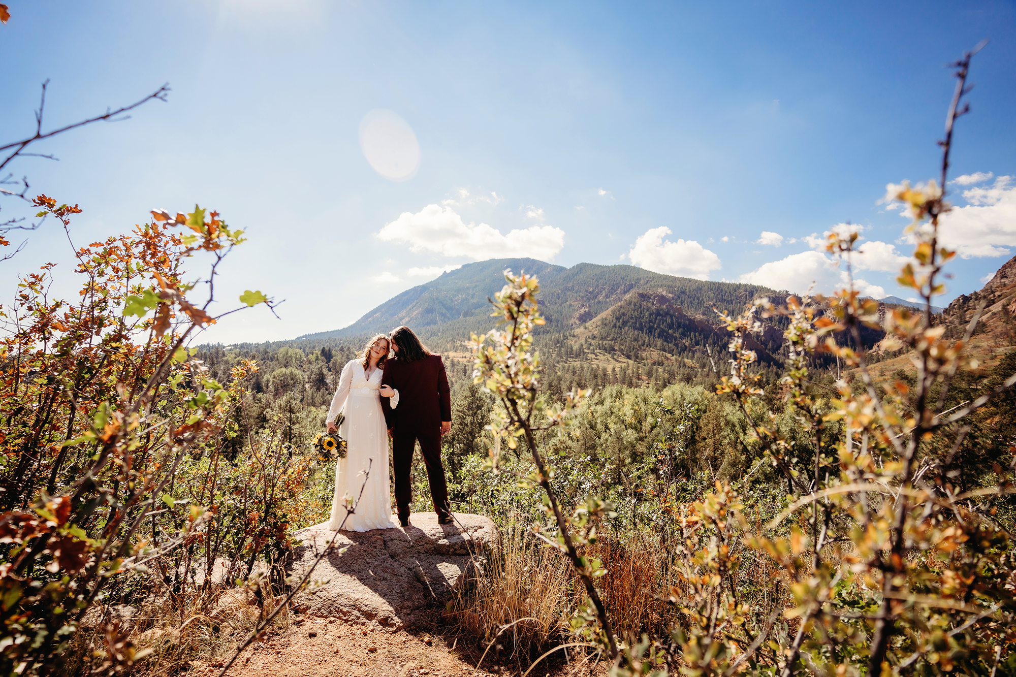 Bride and groom stand on an rock amid mountain foliage on a sunny day in photo captured by Colorado elopement photographers