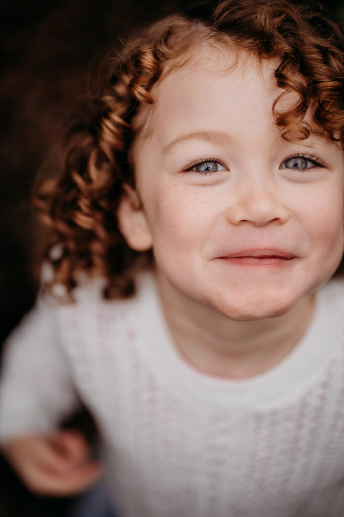 Denver family photographers capture a close up portrait of a little girl with blue eyes and curly brown hair, who smiles slyly at the camera
