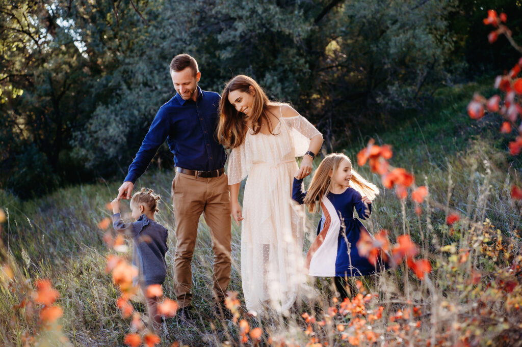A woman in a white maxi dress stands with her partner, a man in blue, while their two kids run through fall foliage by Denver family photographers