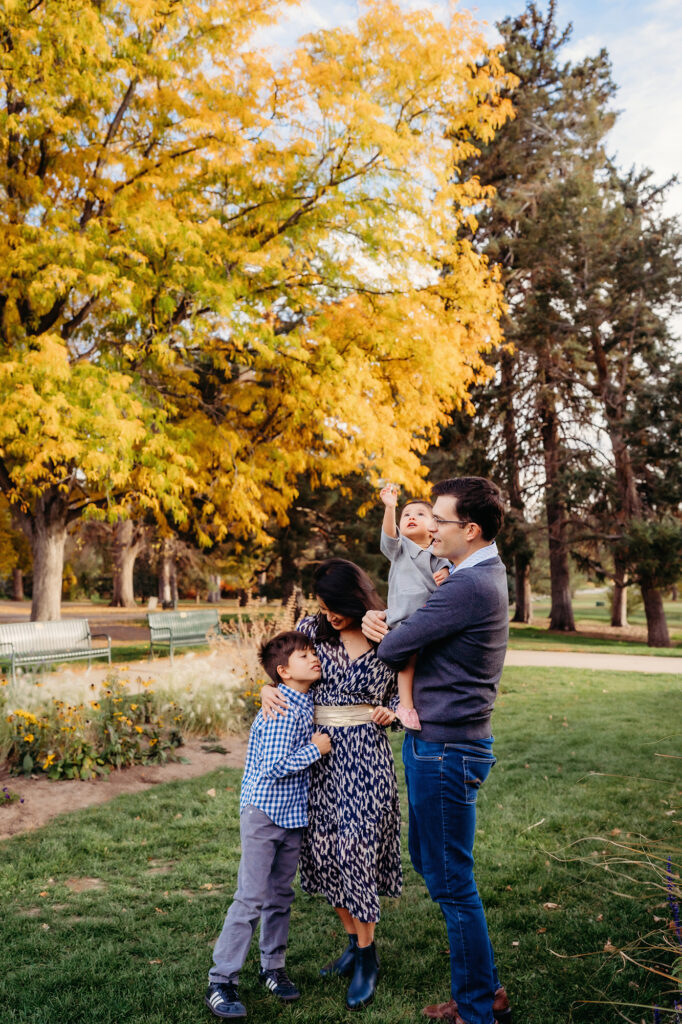 A baby with her family reaches up toward a yellow leaf from an autumn tree during their Denver family photo session with a Denver family photographer