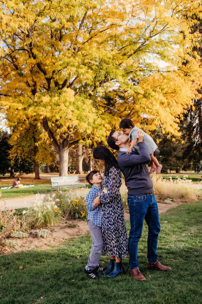 A large yellow tree overshadows a small family of four, who are looking at each other in an unposed moment captured by Denver family photographers