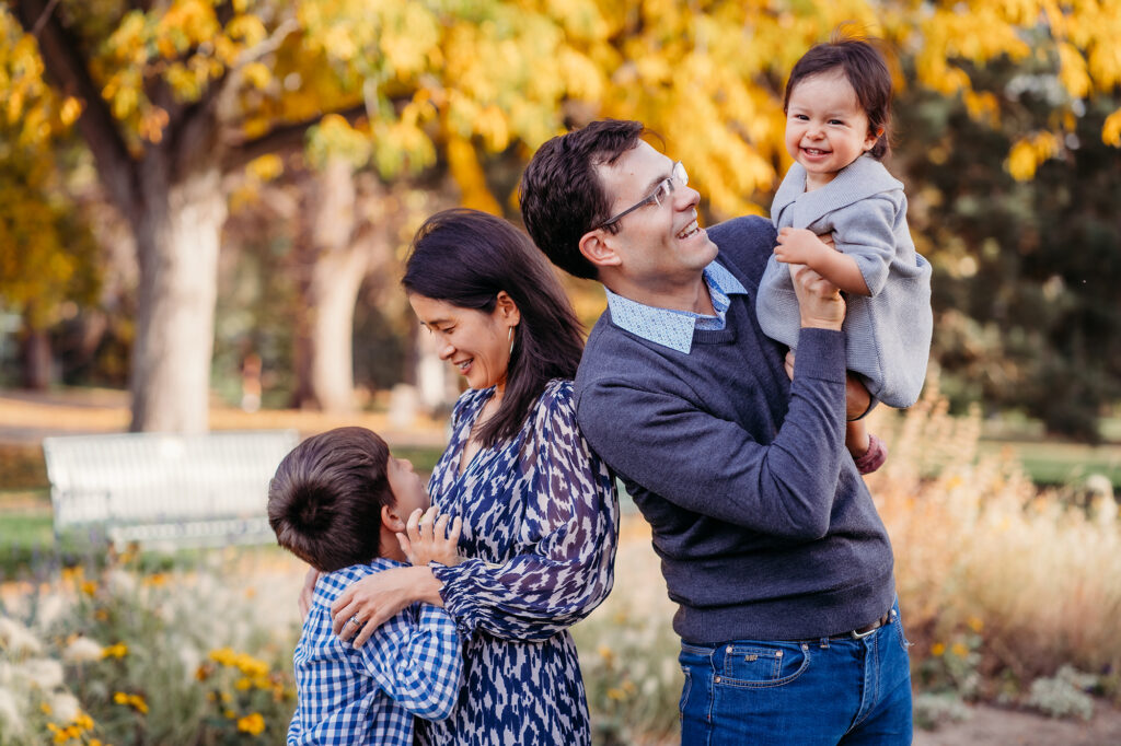 A mother and son look at each other while dad laughs with baby under autumn trees in a photo by Denver family photographers