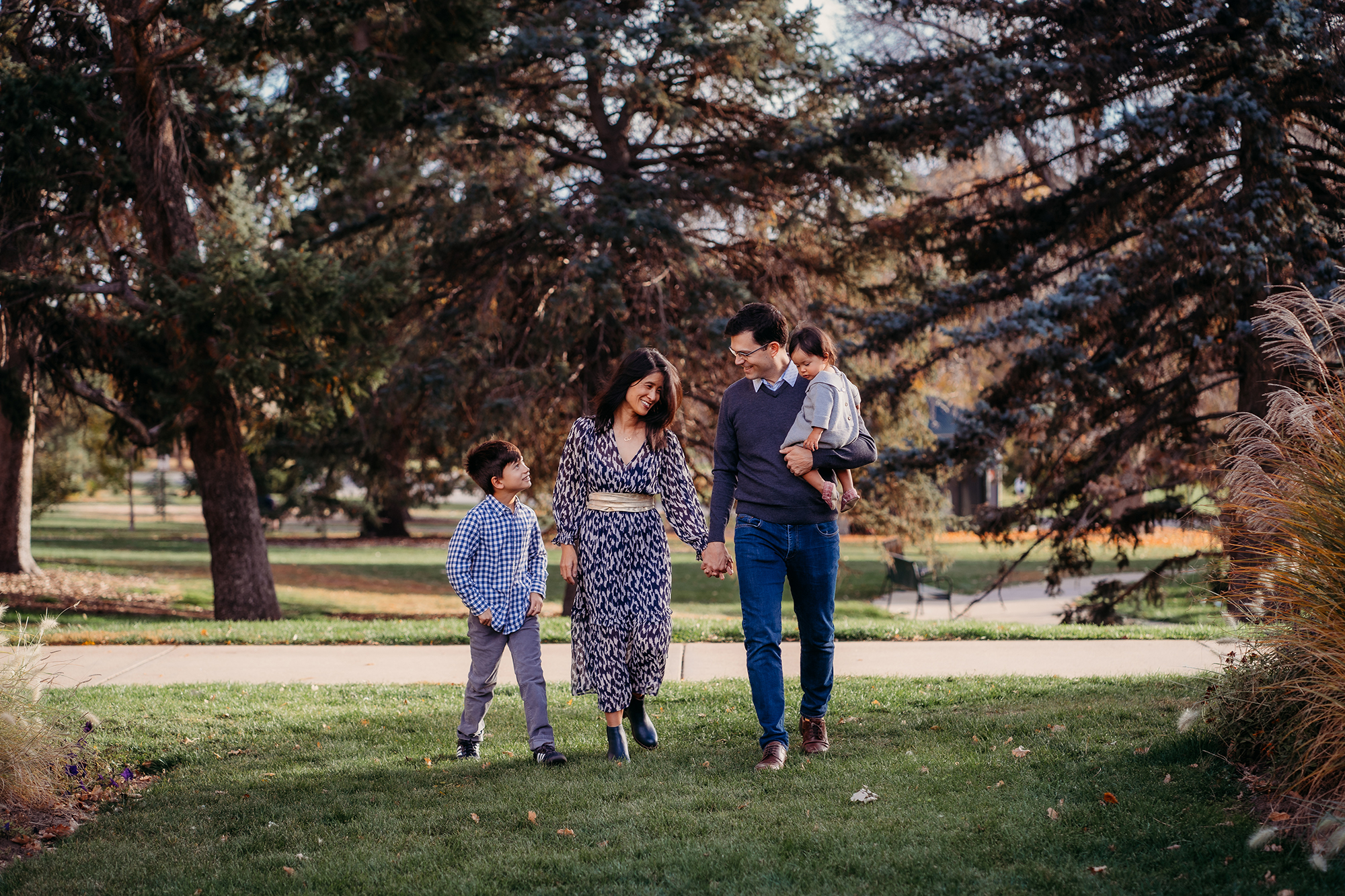 A family walks holding hands under evergreen trees at Cheesman Park in Denver for their Denver family photos with Denver family photographers