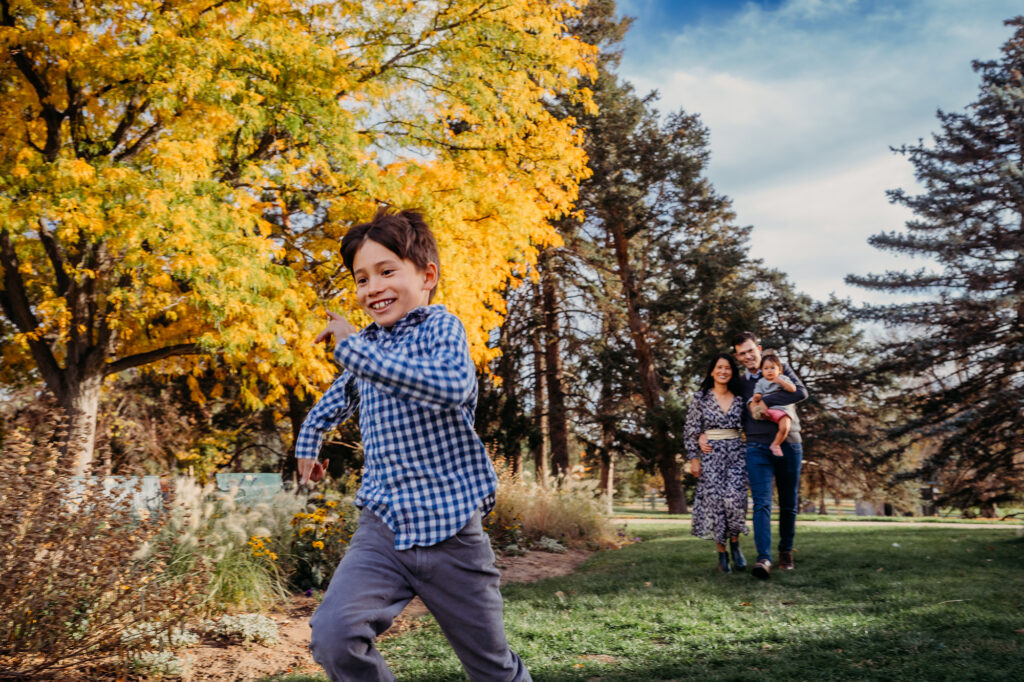 A boy runs past a yellow tree with his family following behind captured by Denver family photographers during their family session in Denver, CO