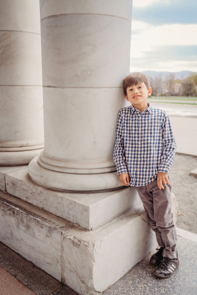 A young boy half-smiles while leaning against a white column in a photo by Denver family photographers