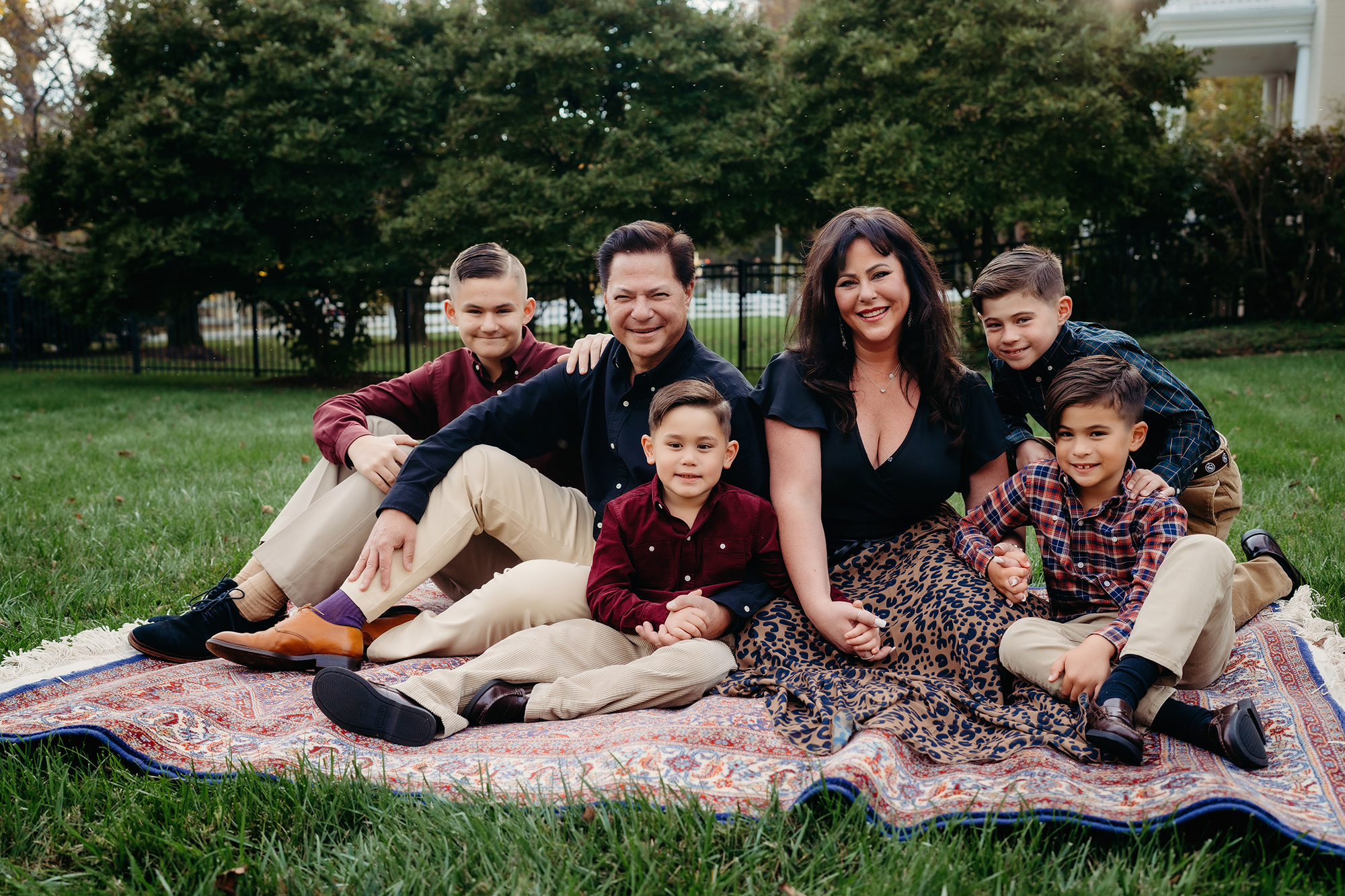 Denver family photographers capture a full family sitting together on a rug in a lush green backyard in Maryland