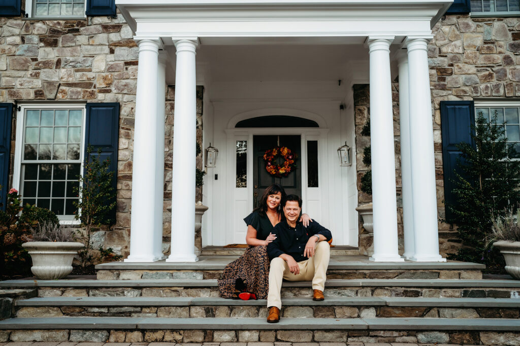 A husband and wife pose on the front step of a colonial-style house for their backyard family session with Denver family photographers