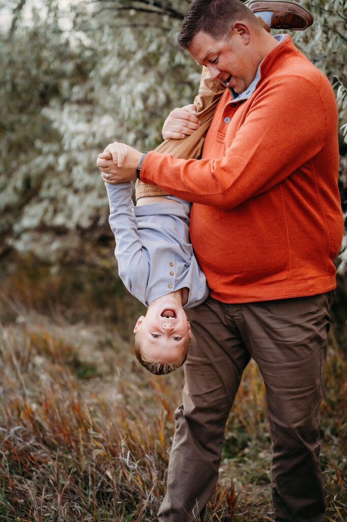 Dad dumps his little boy upside down while boy is laughing in front of fall foliage captured by Denver family photographers