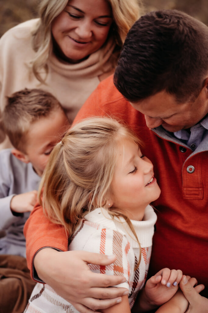 A dad puts his arm around his young daughter in the foreground while mom plays with son in the background of a photo by Denver family photographers