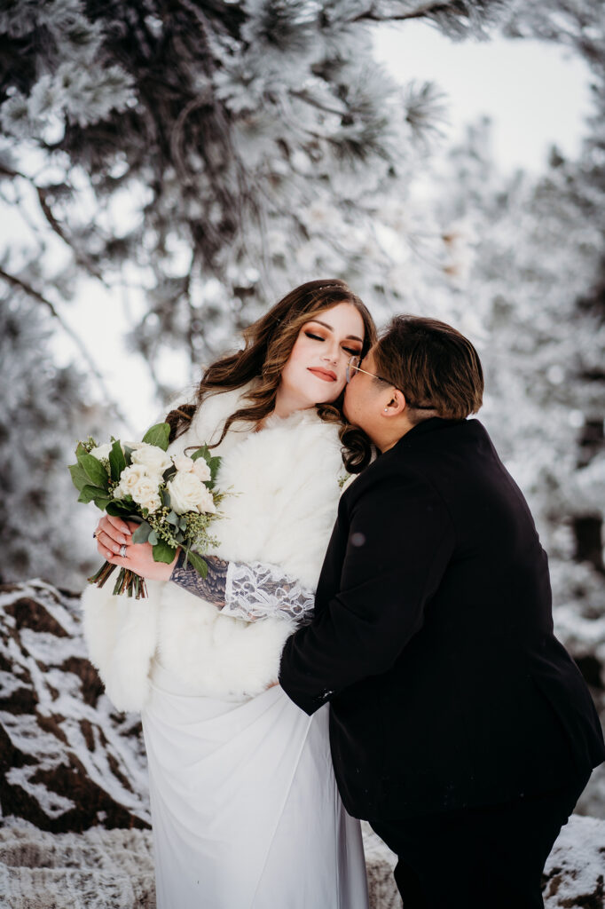 A bride in a suit kisses the cheek of her partner, who wears a white dress and holds a bouquet in this portrait by Colorado elopement photographer