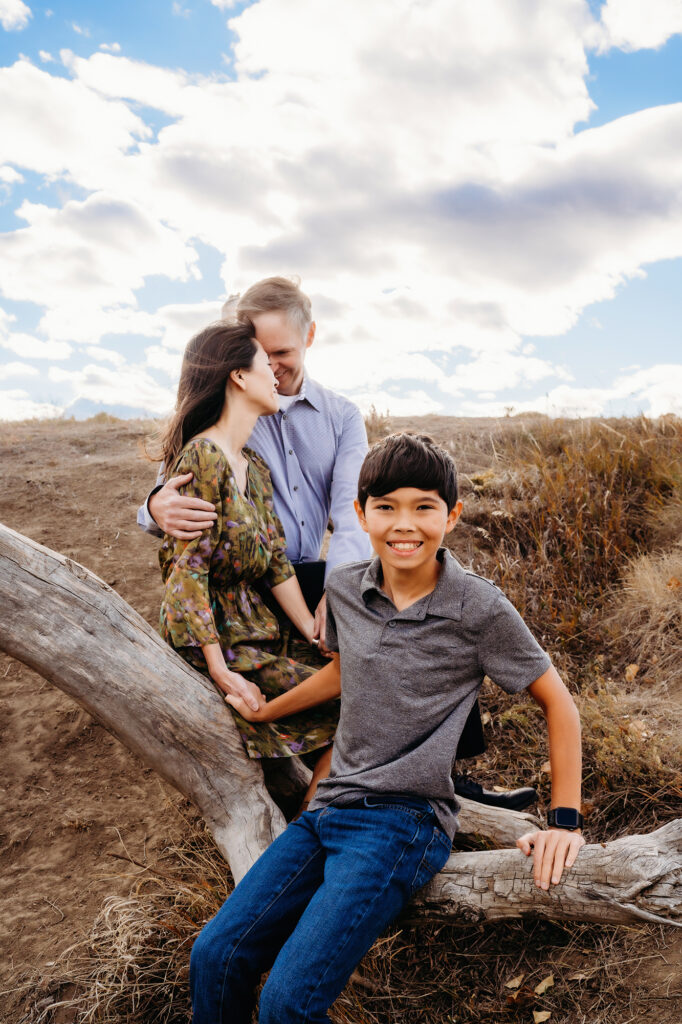 A father and mother embrace while their son sits on a large branch of driftwood at Standley Lake captured by Denver family photographers