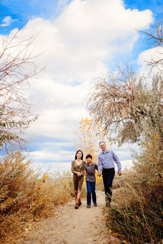 Under a cloudy blue sky, a mom, son, and dad hold hands and walk along a beach path captured by Denver family photographers