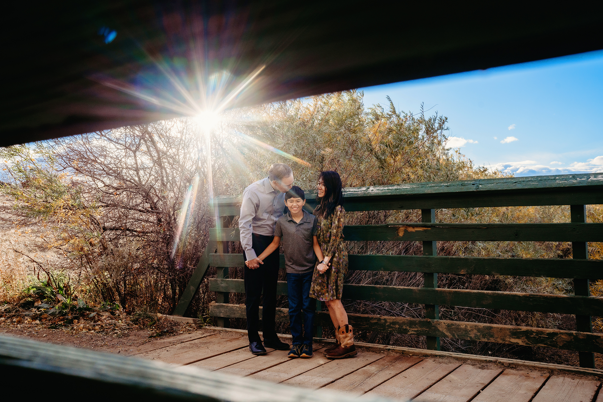 Denver family photographers capture a solar flare over a family of three holding hands on a green bridge at Standley Lake, CO