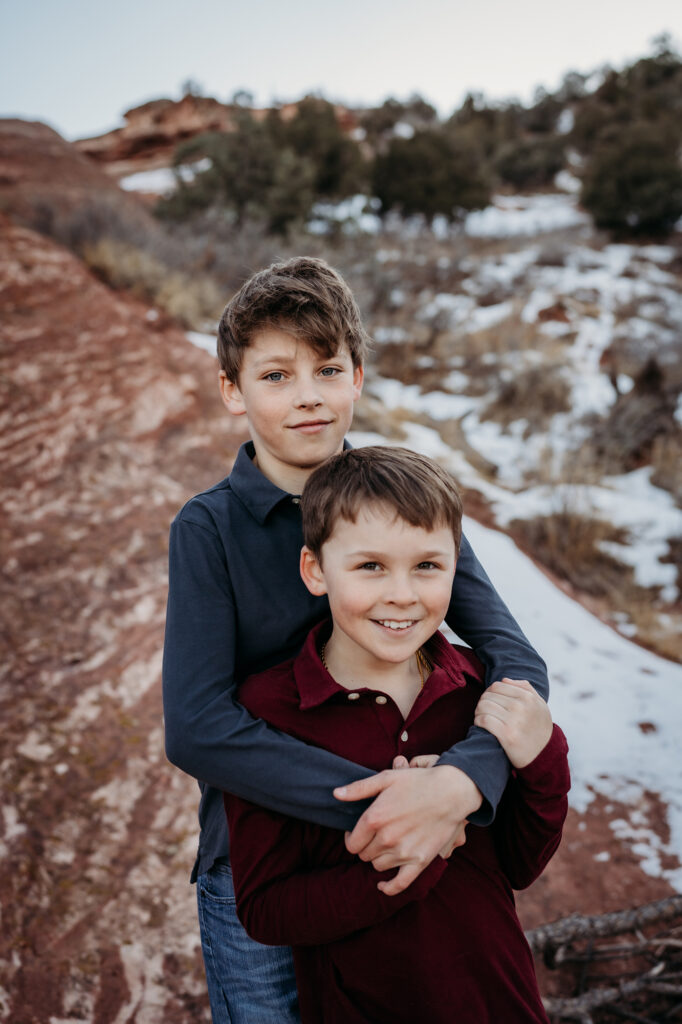 denver family photographers captures brother hugging each other in the red rocks with snow surrounding them for their winter family photos