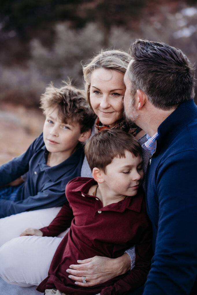 mother sitting with her arms around her two sons as the wind blows her hair out of her face and her husband leans in to kiss her forehead photographed by denver family photographers