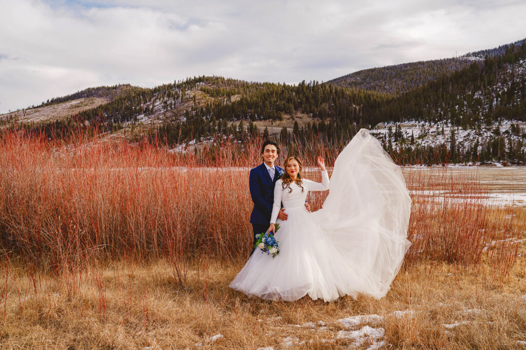 outdoor elopement with Colorado elopement photographer capturing bride trowing her dress in the wind as her groom embraces her waist with the mountains in the distance