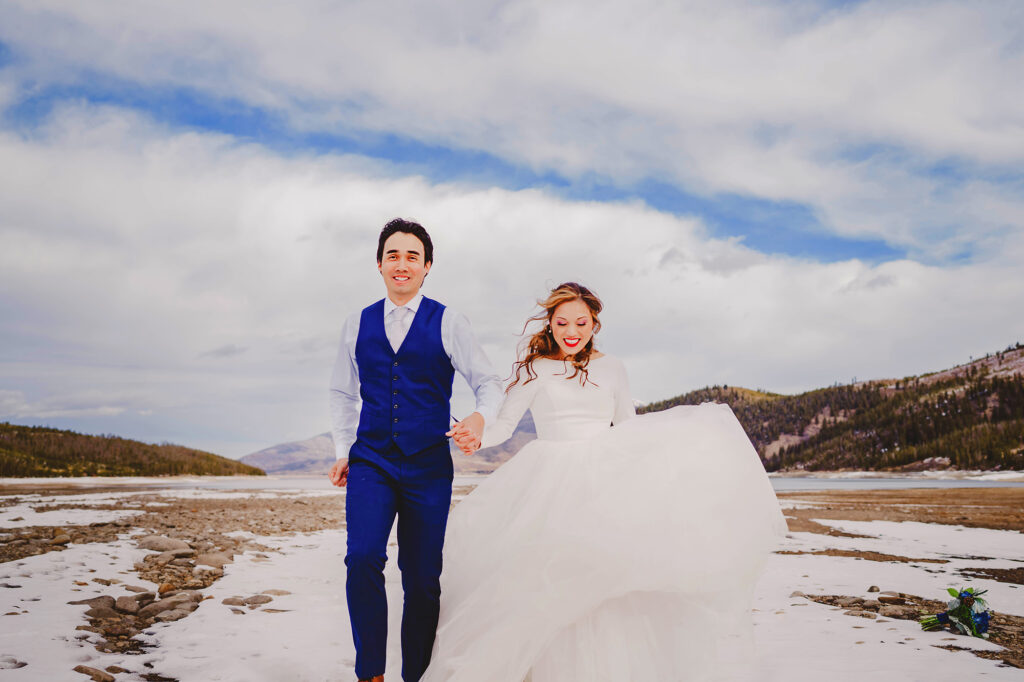 Colorado elopement photographs captures bride and groom holding hands and running through a snowy field in the Rockies for their destination elopement