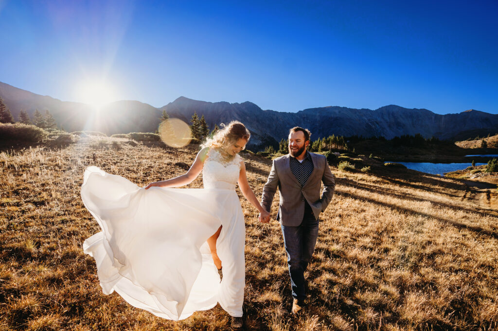 sunset wedding pictures with bride and groom holding hands and walking through a field together with the mountains in the distance as the sun shines right over them