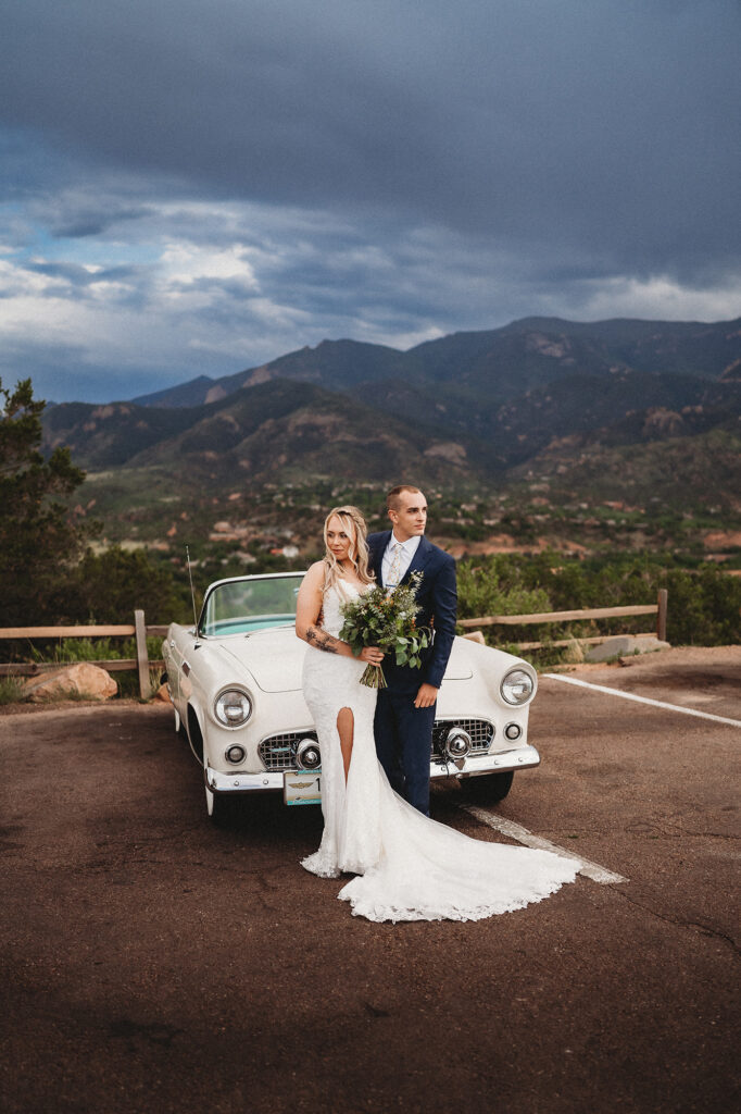 rainy day mountain wedding captured by Colorado elopement photographer with bride and groom posing in front of a white vintage car while with dark storm clouds in the distance