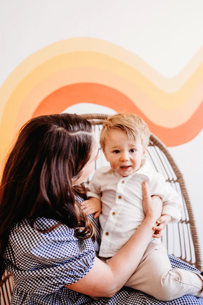 young mother holding her toddler son and playing as he laughs and reaches for the camera with a groovy wall and decor behind them 