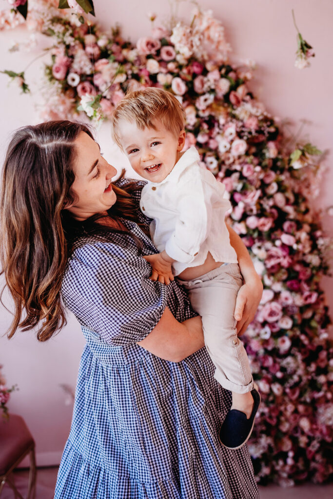 mother son photoshoot with mom in a blue dress holding her toddler son and laughing with her with a pink flower backdrop captured by Denver family Photographers