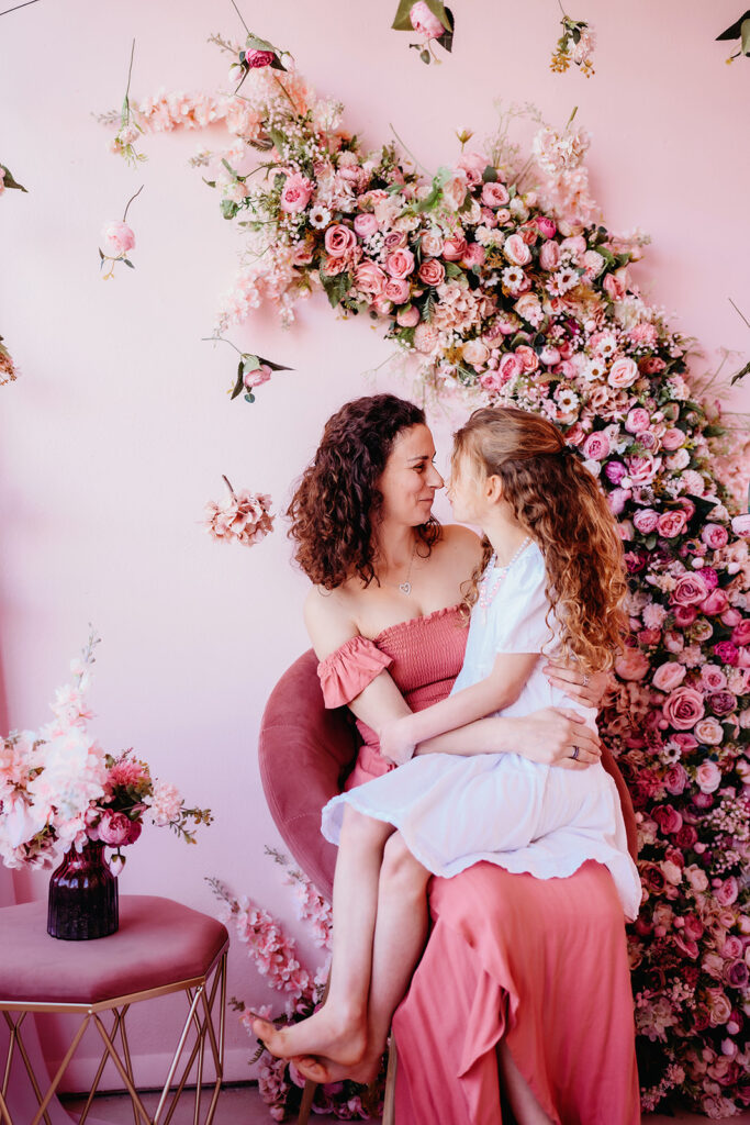 mommy and me photos for mothers day gift ideas with mom sitting on a stool with a pink backdrop and talking with her young daughter who is sitting in her lap