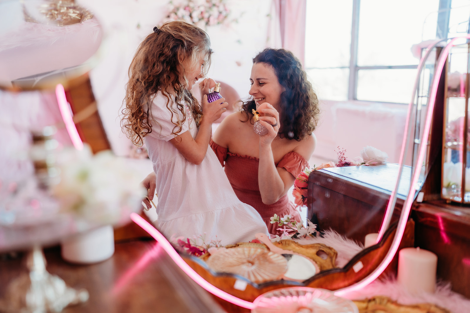 mother daughter photoshoot with mother kneeling next to her young child as they hold perfume bottles together and smile while in front of a vanity mirror captured by Denver family Photographers