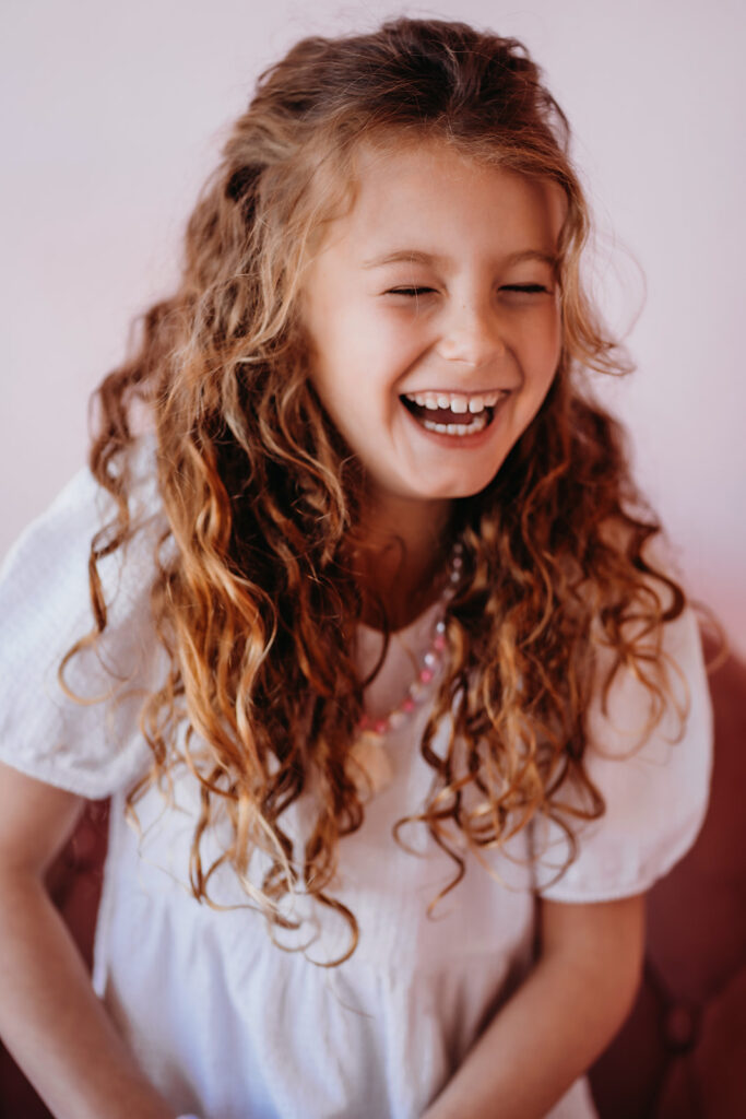 headshot of little girl with curly hair laughing as she wears a white dress sitting on a pink chair captured by Denver family Photographers