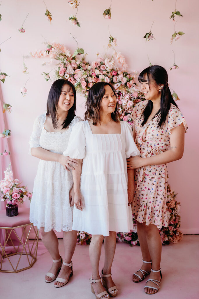daughters standing side by side with their mother in a studio decorated for a mothers day shoot with pink walls and floral arrangements 