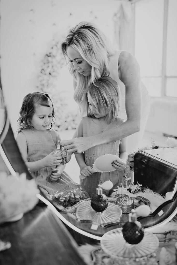 Denver family Photographers captures mother with her two young daughters at a vanity putting on perfume and makeup with flowers in the distance
