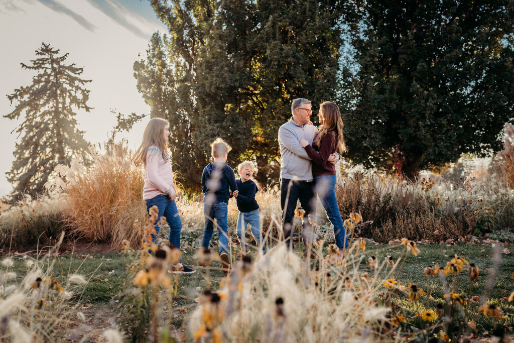 fall family photos with denver family photographers with children running around their parents as they stand in a field together with a grove of trees to the right