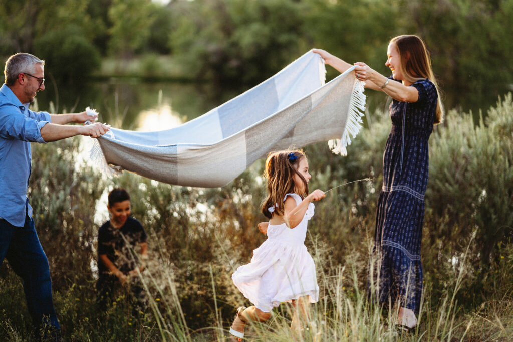 Denver family photographers photographs candid image of little children playing with their parents as the adults wave a blanket in the air next to some shrubs near a river