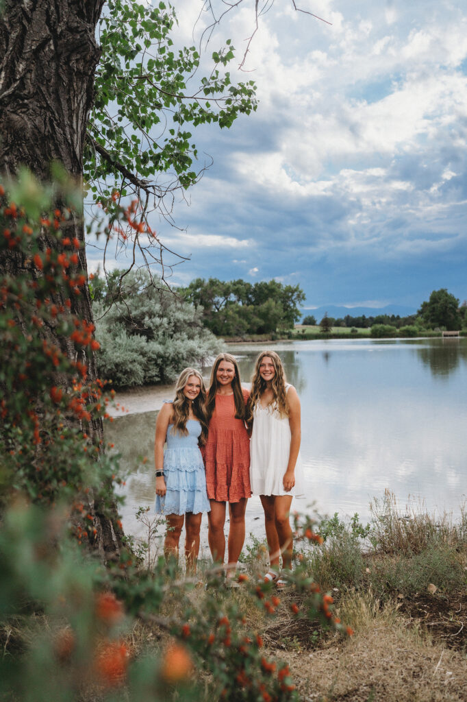 denver family photographers photographs three sisters in dresses standing on the edge of the lake with a cloudy sky in the distance