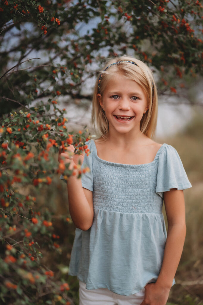 denver family photographers captures headshot of little girl in a blue top standing next to a bush with orange flowers smiling