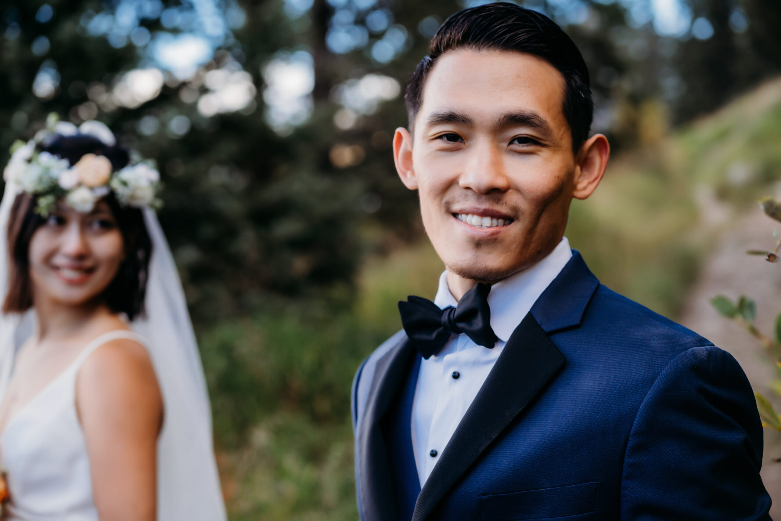 asain groom in a blue suit with a black bowtie smiles as his bride stands in the distance looking at him for their denver wedding in a forest