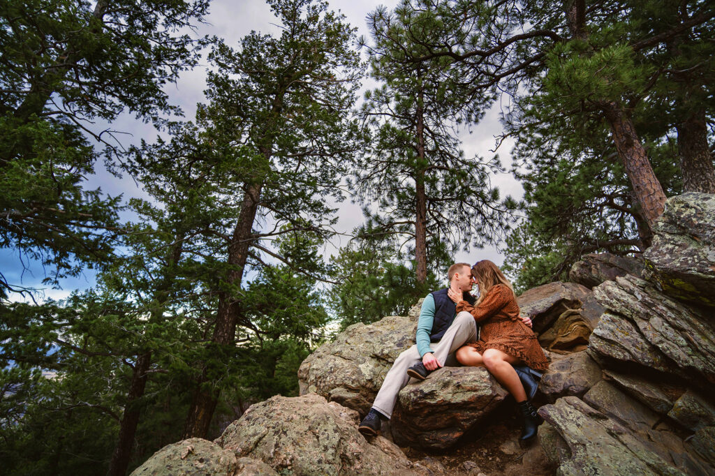 colorado elopement photographer captures outdoor engagement photos with man and woman sitting on a rock in the woods and kissing for couple photoshoot outfits