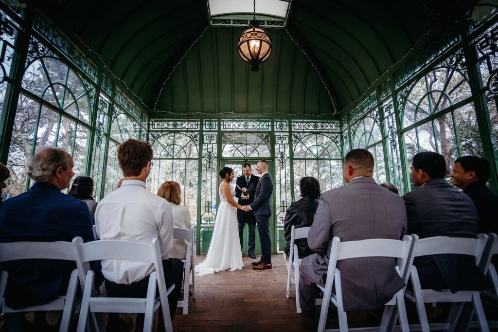 bride and groom holding hands in side of a greenhouse with their guests sitting and watching them as they get married 