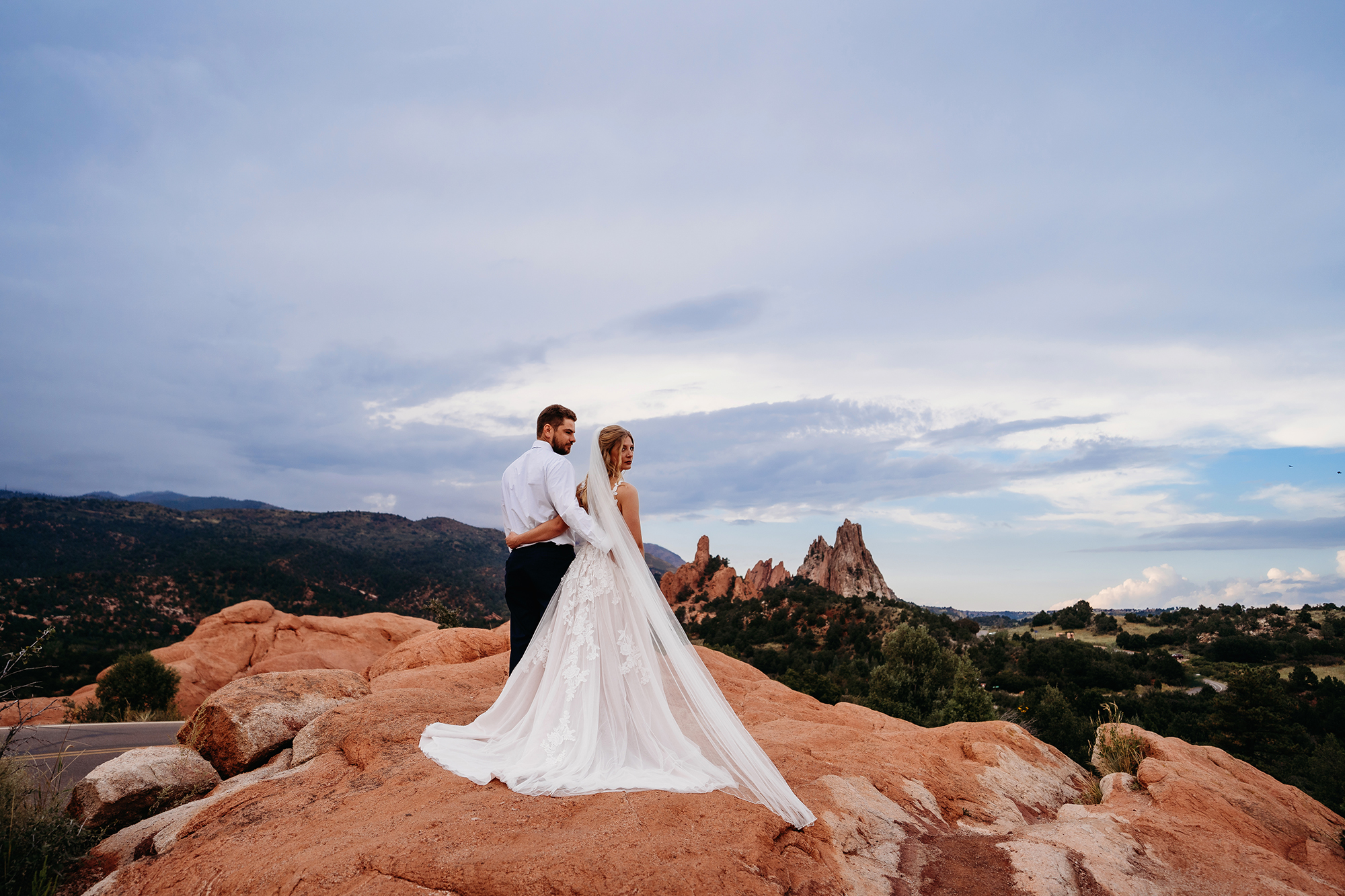 bride and groom standing on a mountain top in the red rocks and embracing one another while looking out to the valley of trees and shrubs for their colorado elopement
