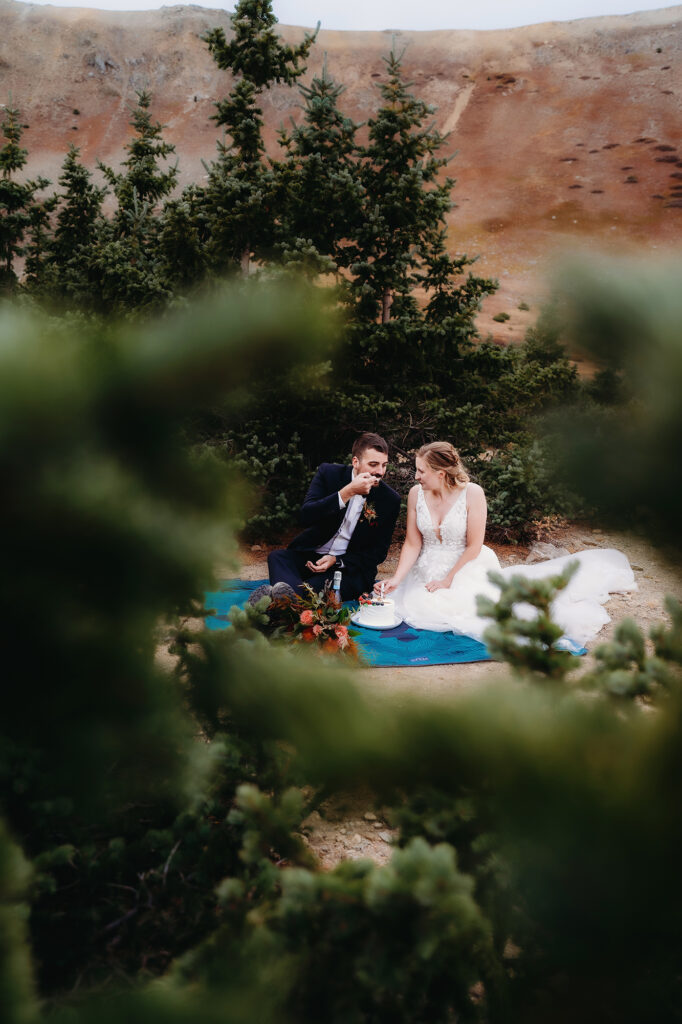 bride and groom sitting on a teal blanket in the woods eating cake together for their colorado elopement