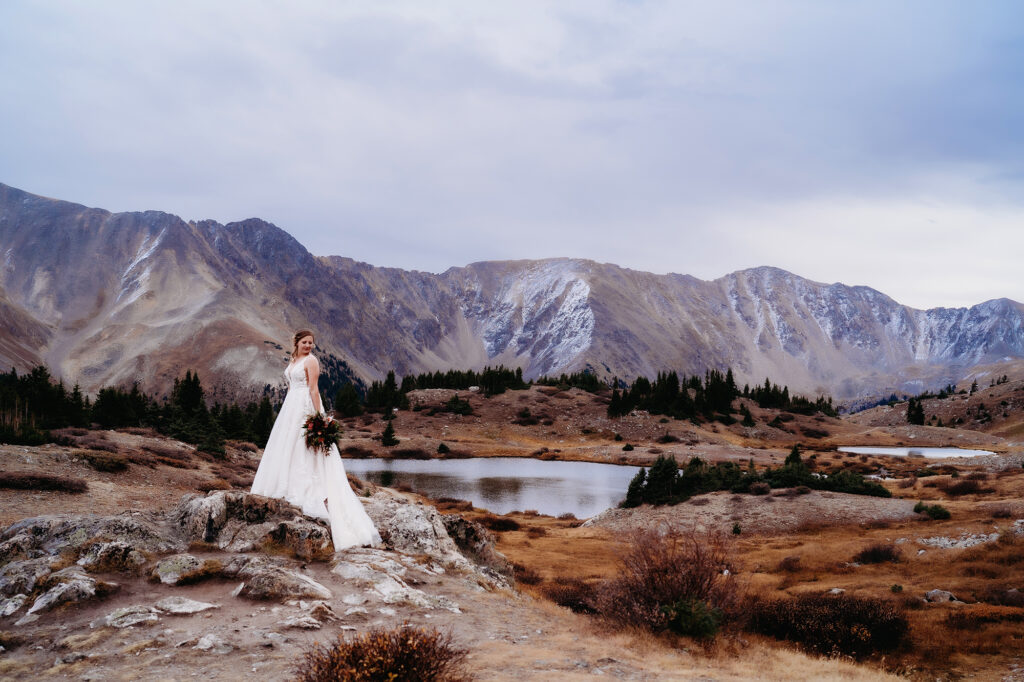 bride standing on a boulder holding her bouquet to her side and looking over her shoulder with a lake behind her and mountains in the distance photographed by colorado elopement photographer