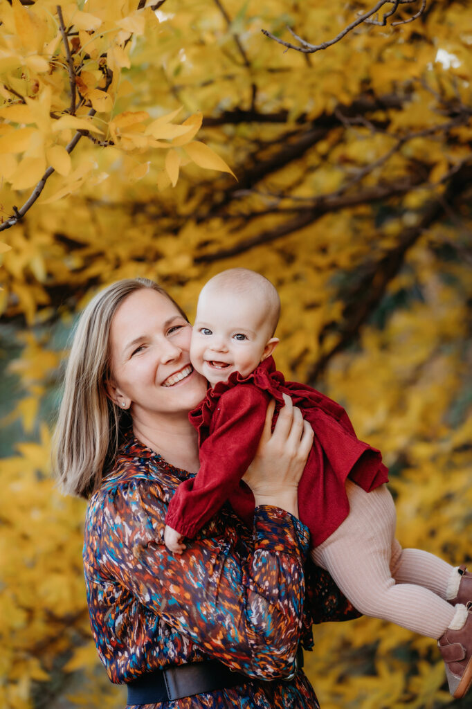 mother holding baby next to her shoulder and pressing their cheeks together as they smile in a forest with autumn leaves