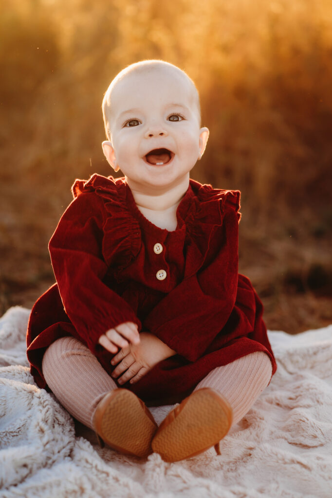 Denver family photos with baby girl in a red dress sitting on a blanket and laughing while the sun sets in a golden autumn field 