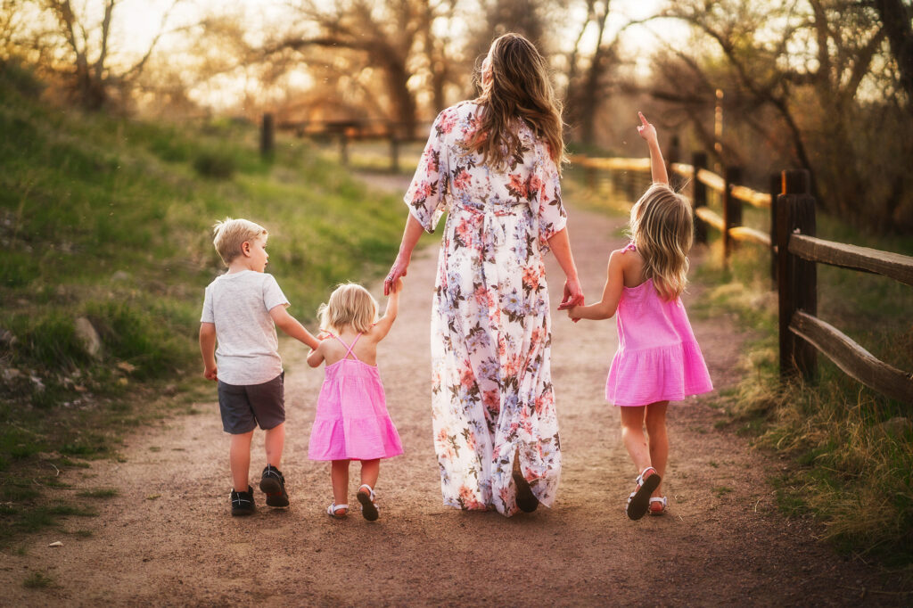 single mom holding hands with her three children and walking down a dirt path while the sun sets through the trees in a local part