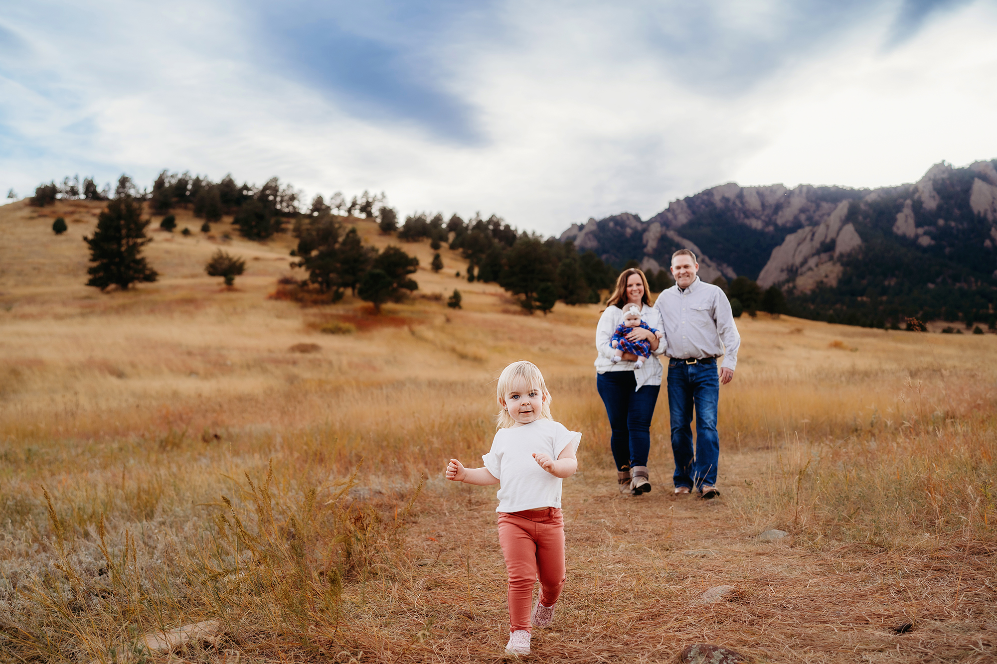 parents and a baby standing together in an open field in the fall with mountains and a forest behind them, while toddler runs up the trail ahead of them