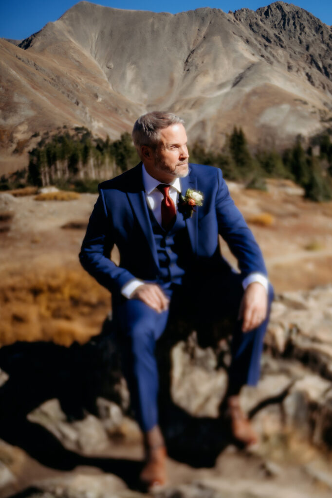 Colorado Elopement Photographer captures groom sitting on rock wearing blue suit looking off into distance