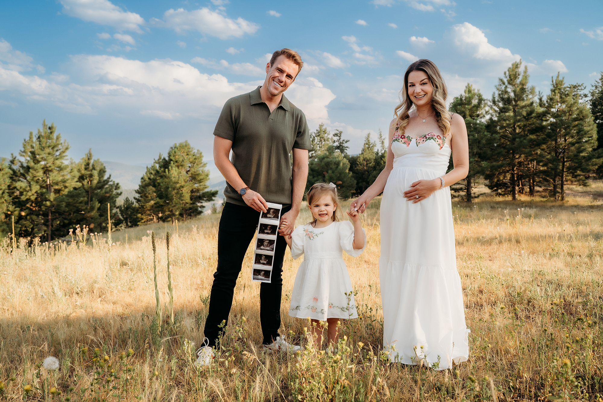 Denver Family Photographer captures young family holding hands in field while showing ultrasound photos