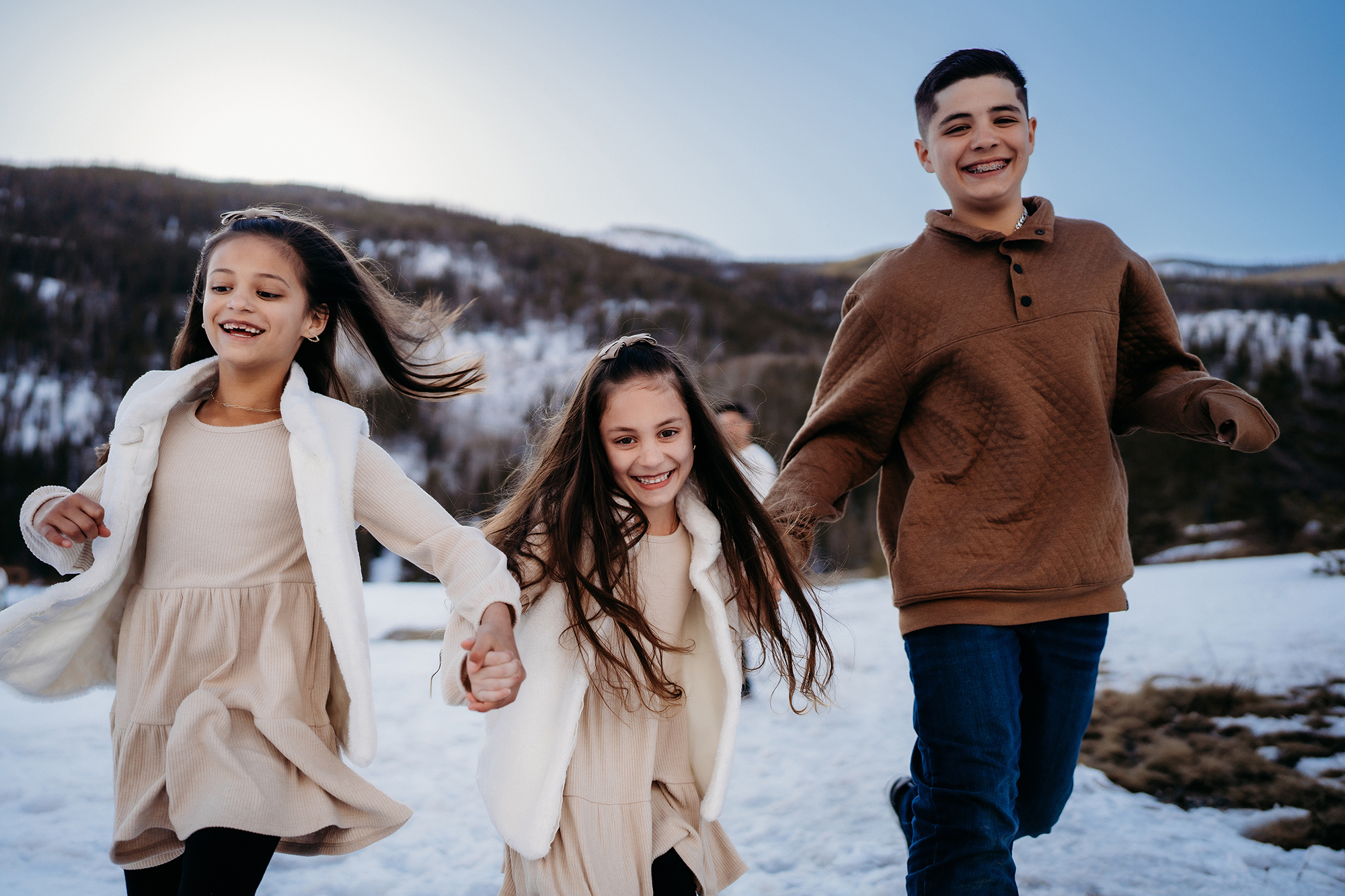 Siblings running hand in hand through snowy mountains, wearing neutral winter outfits.
